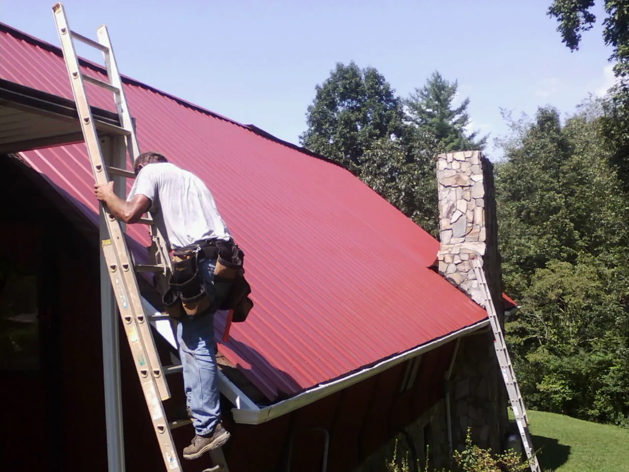 Roofer on a ladder beside a red metal roof and stone chimney in Ellinwood